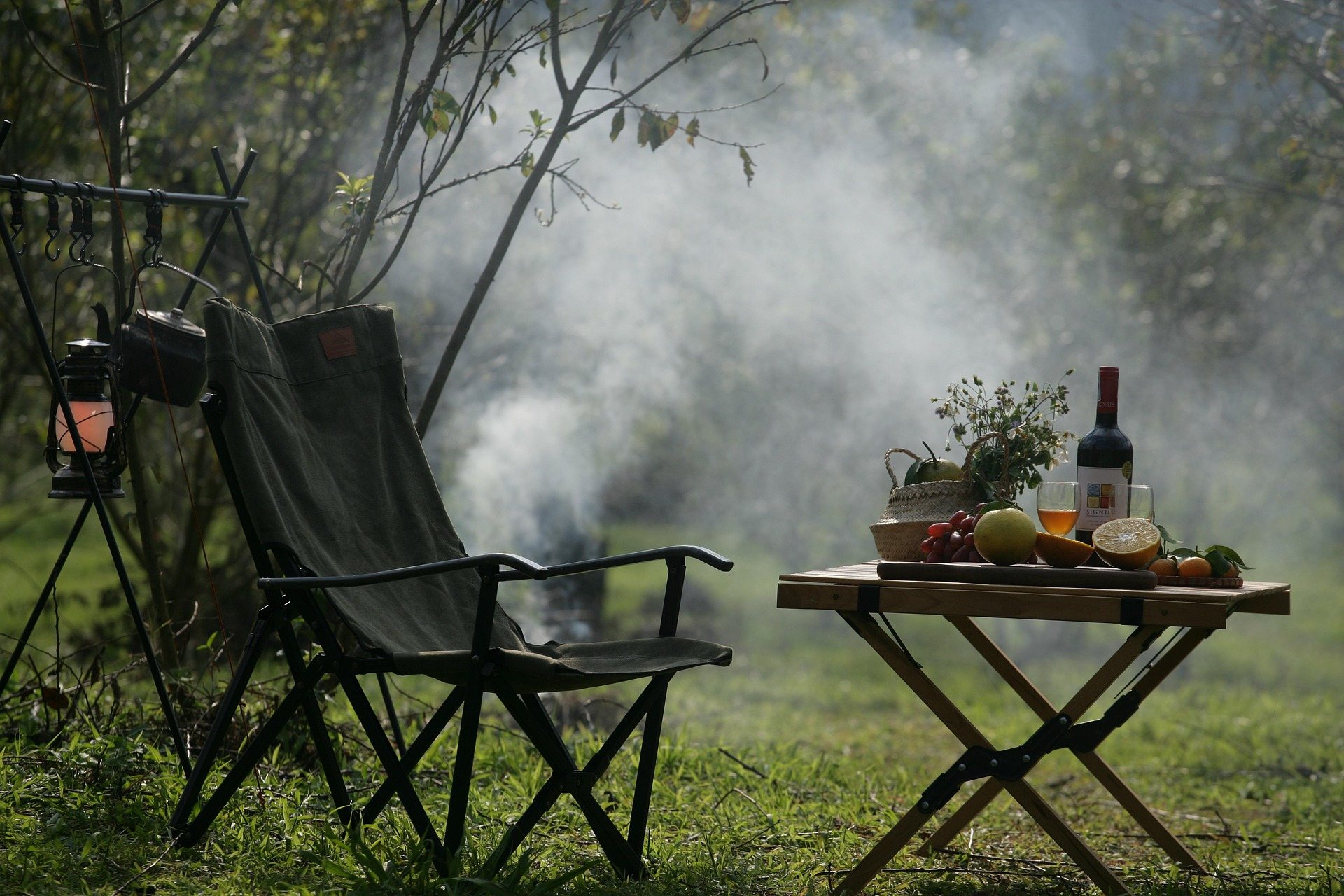 Two camp chairs and a folding table with food and drink set up beside woods in morning mist