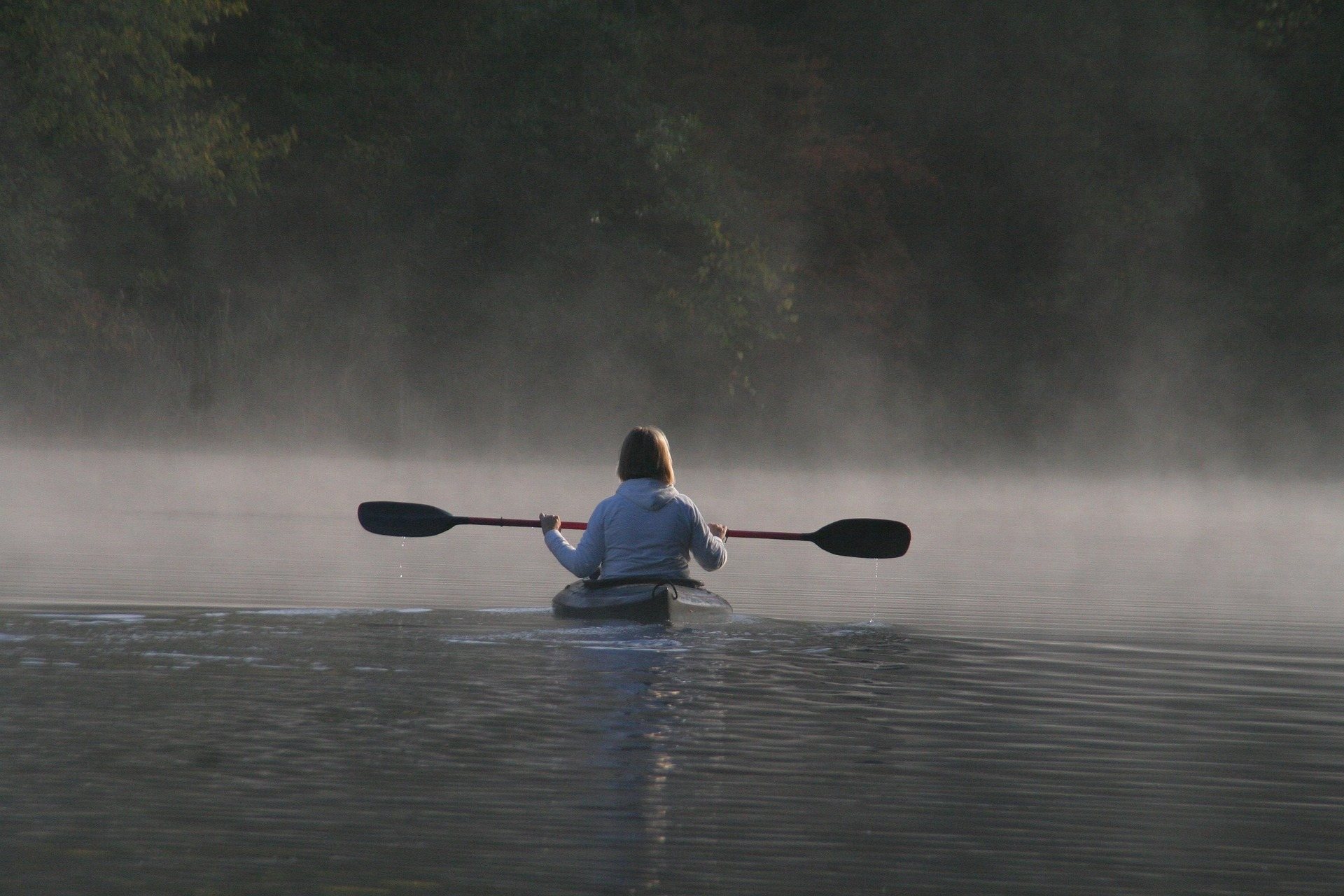 A lone kayaker paddling across still water into low morning mist