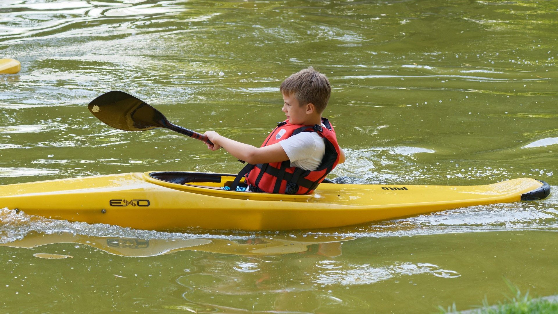 A child in a life jacket paddling a small yellow kayak on calm water