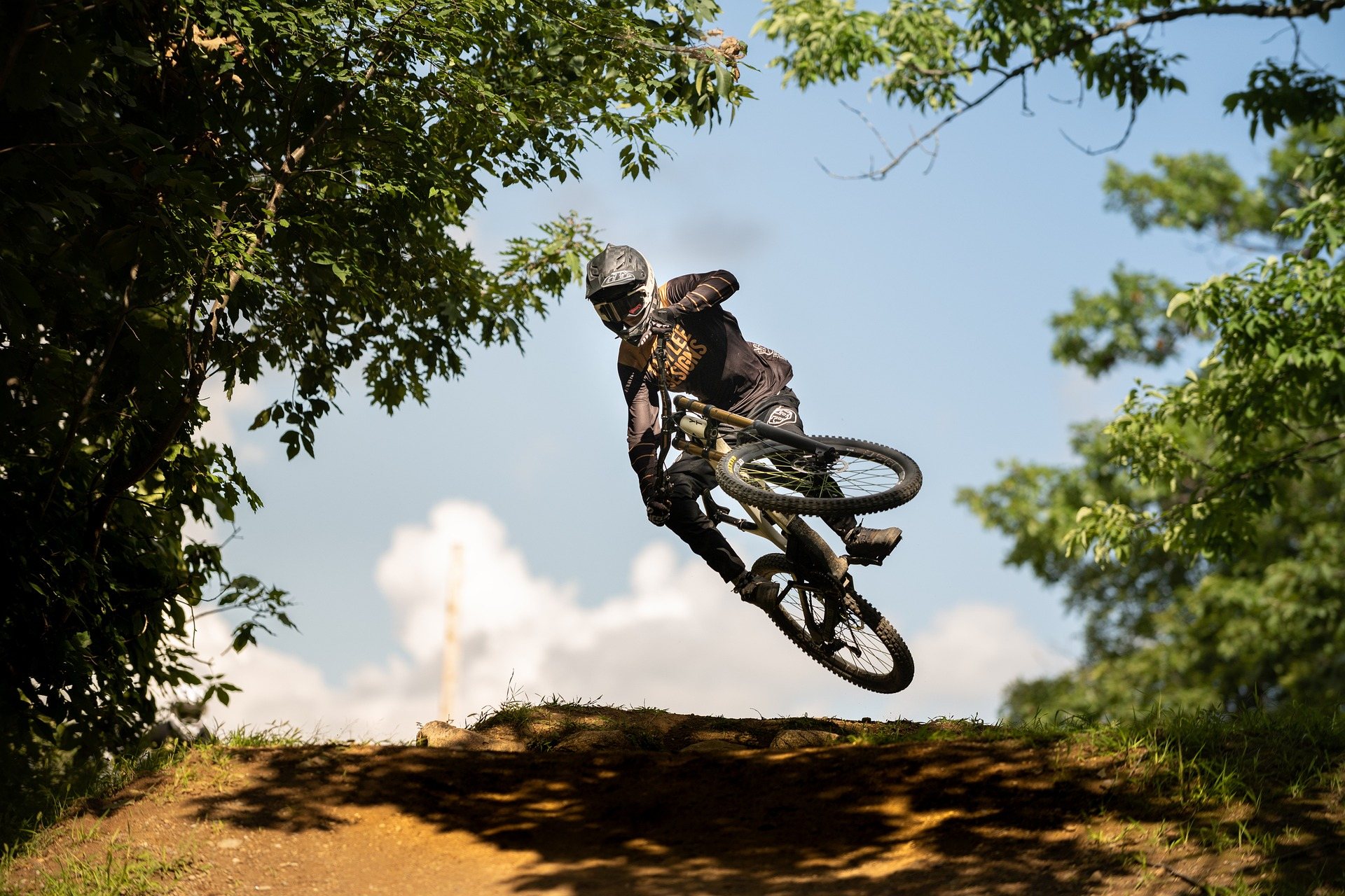 A mountain biker airborne off a dirt jump against a blue sky, framed by summer trees