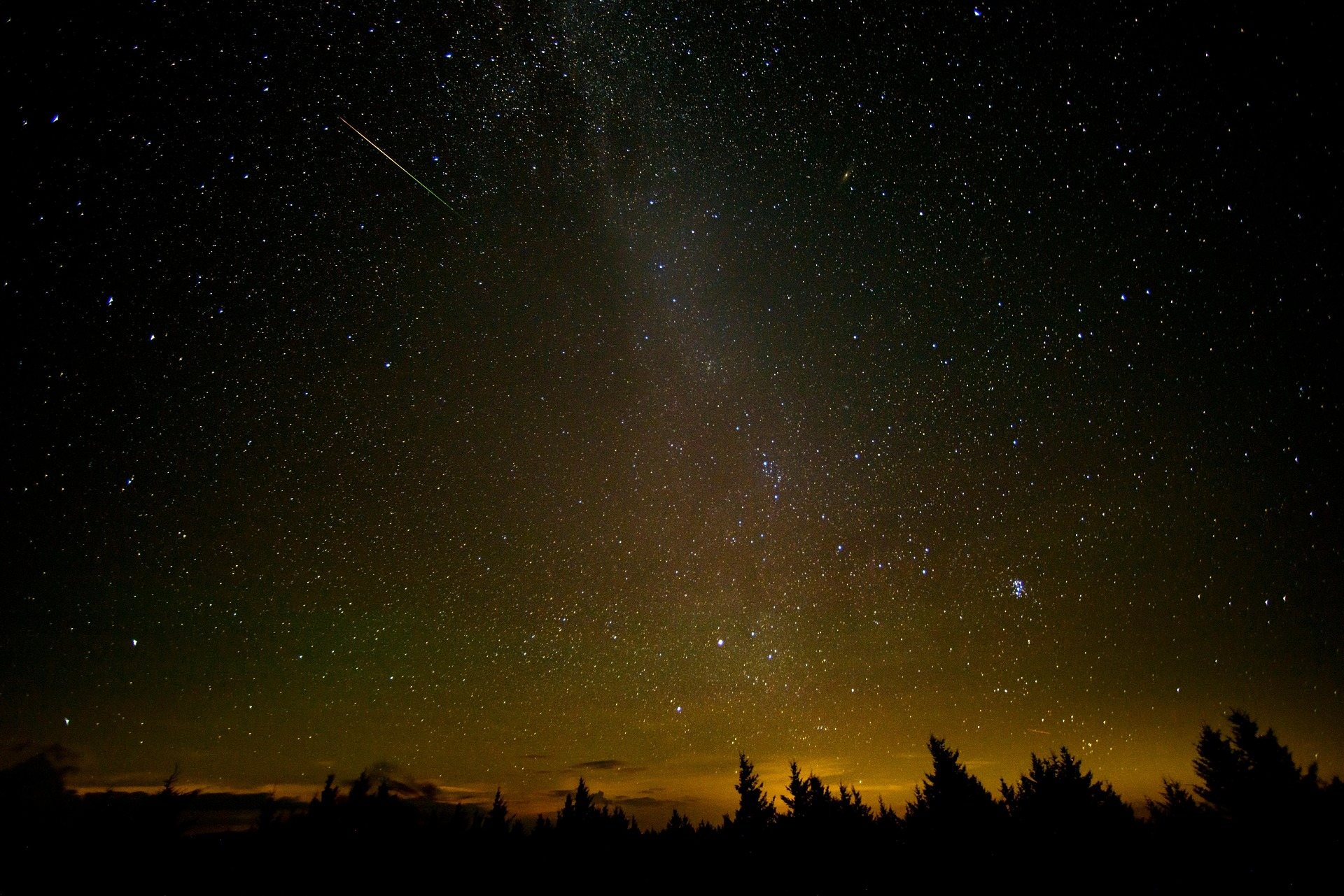 The Milky Way and a meteor streak over a dark treeline, with faint orange light on the horizon