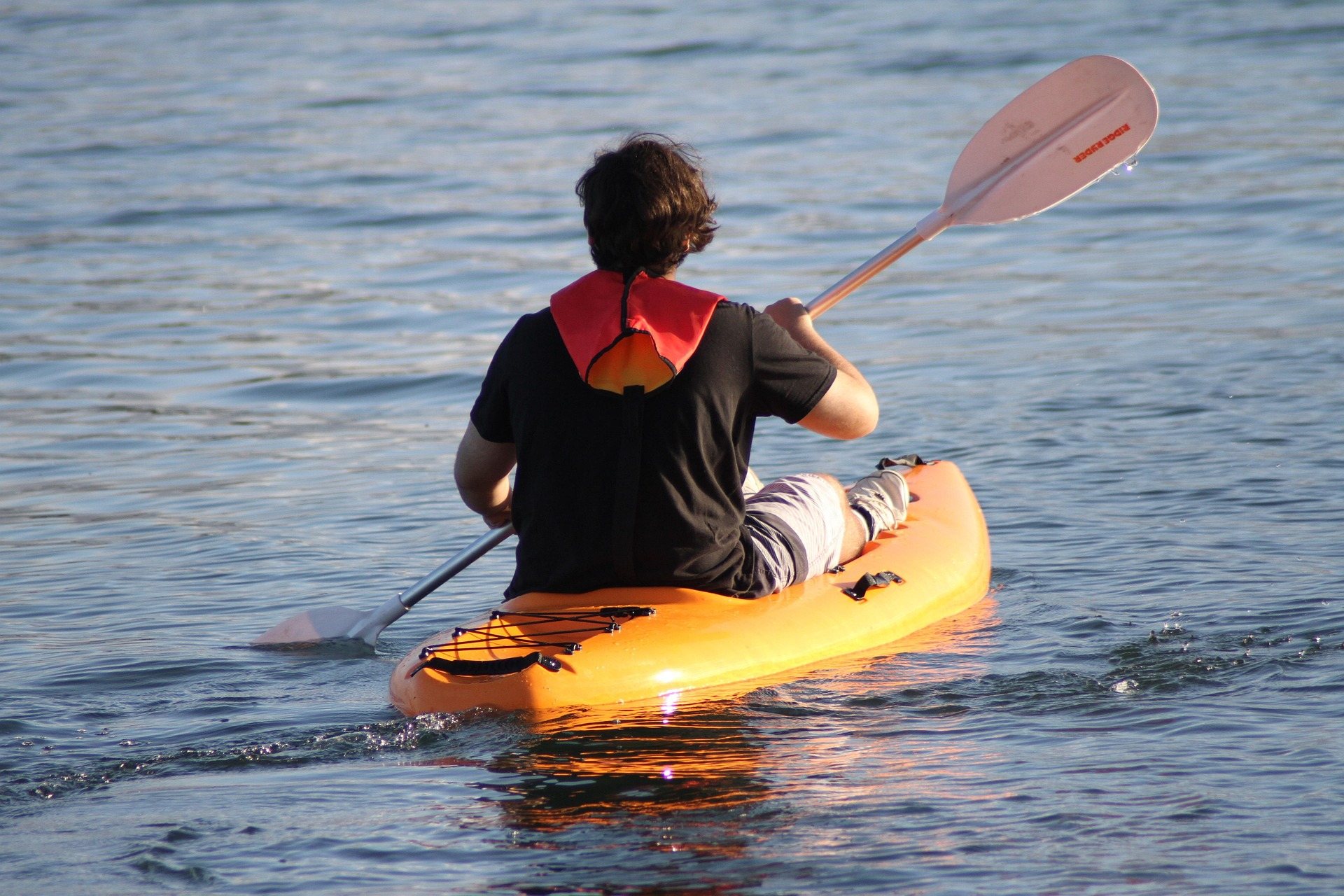 A paddler seen from behind working a sit-on-top kayak across open flat water