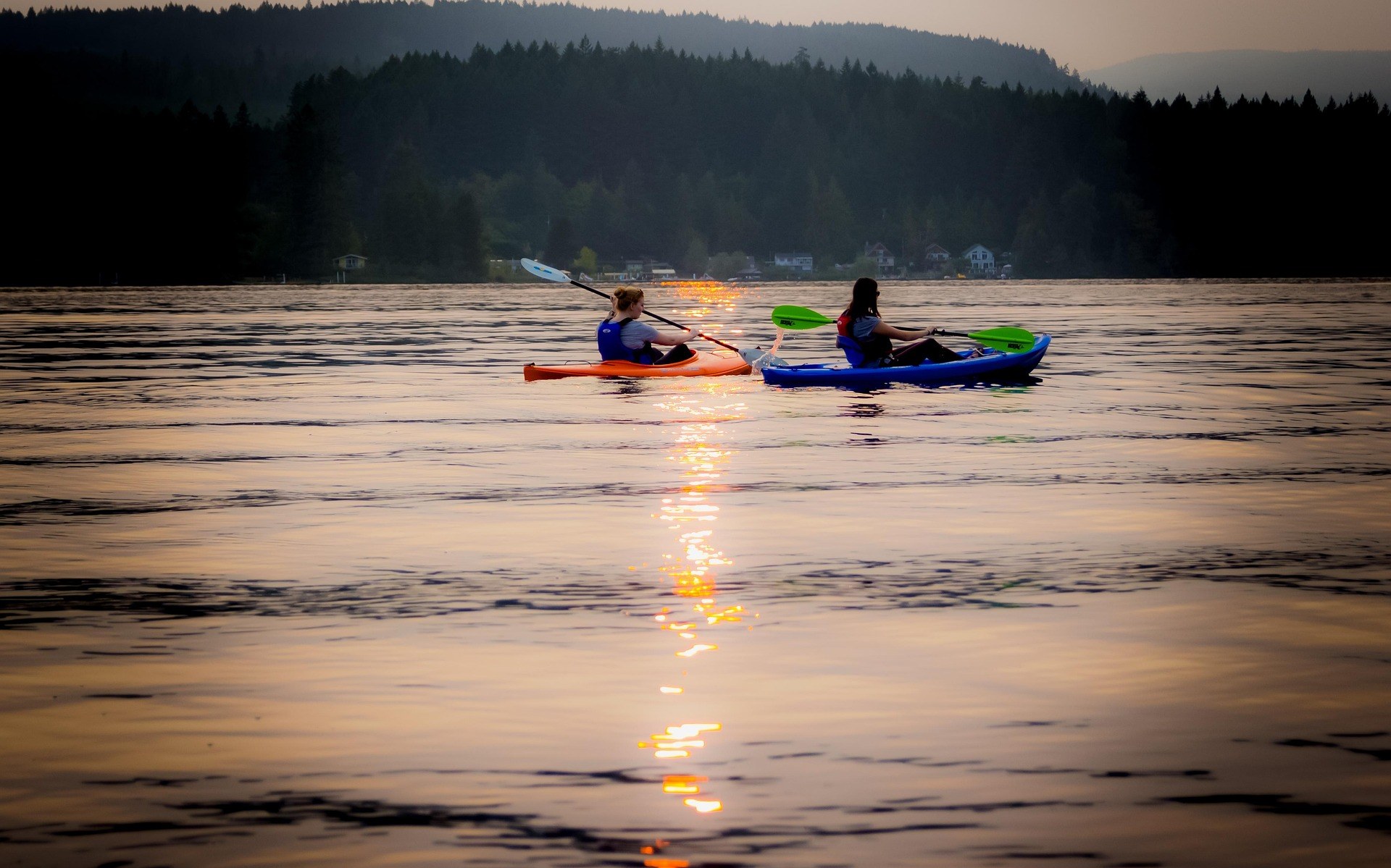 Two kayakers paddling across a still lake at sunset with forested hills on the far shore