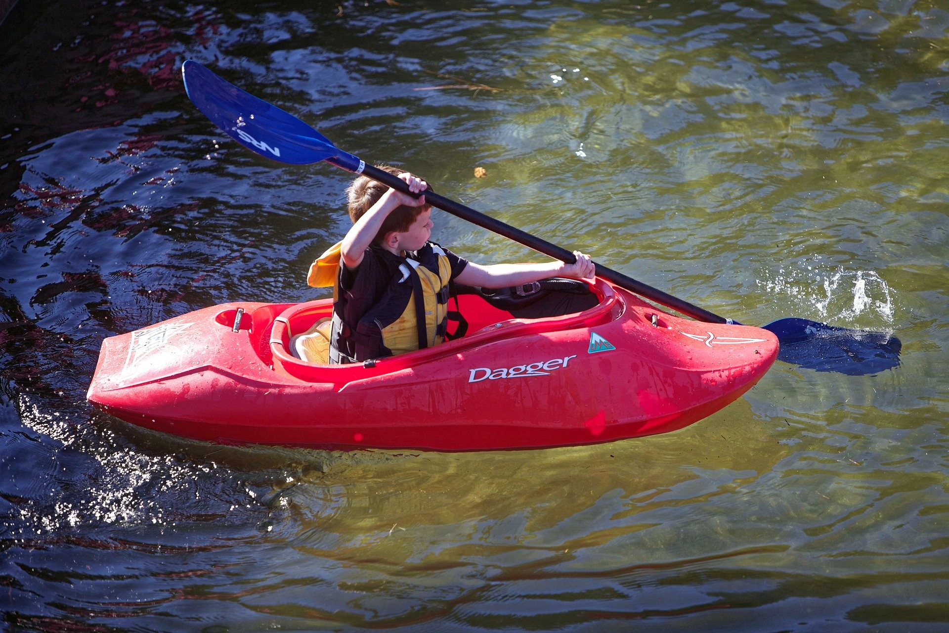 A kayaker in a red boat driving through whitewater, paddle blade in the foam and spray around the bow.